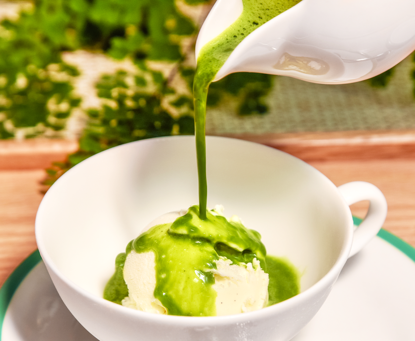 A matcha blend in a white bowl on a white table next to a spoonful of matcha and a silver bulk matcha bag